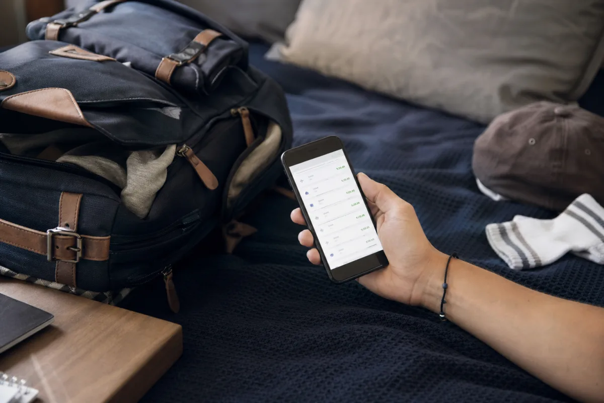 Mobilskärm med en flygprisjämförelse-app öppen bredvid en packad ryggsäck